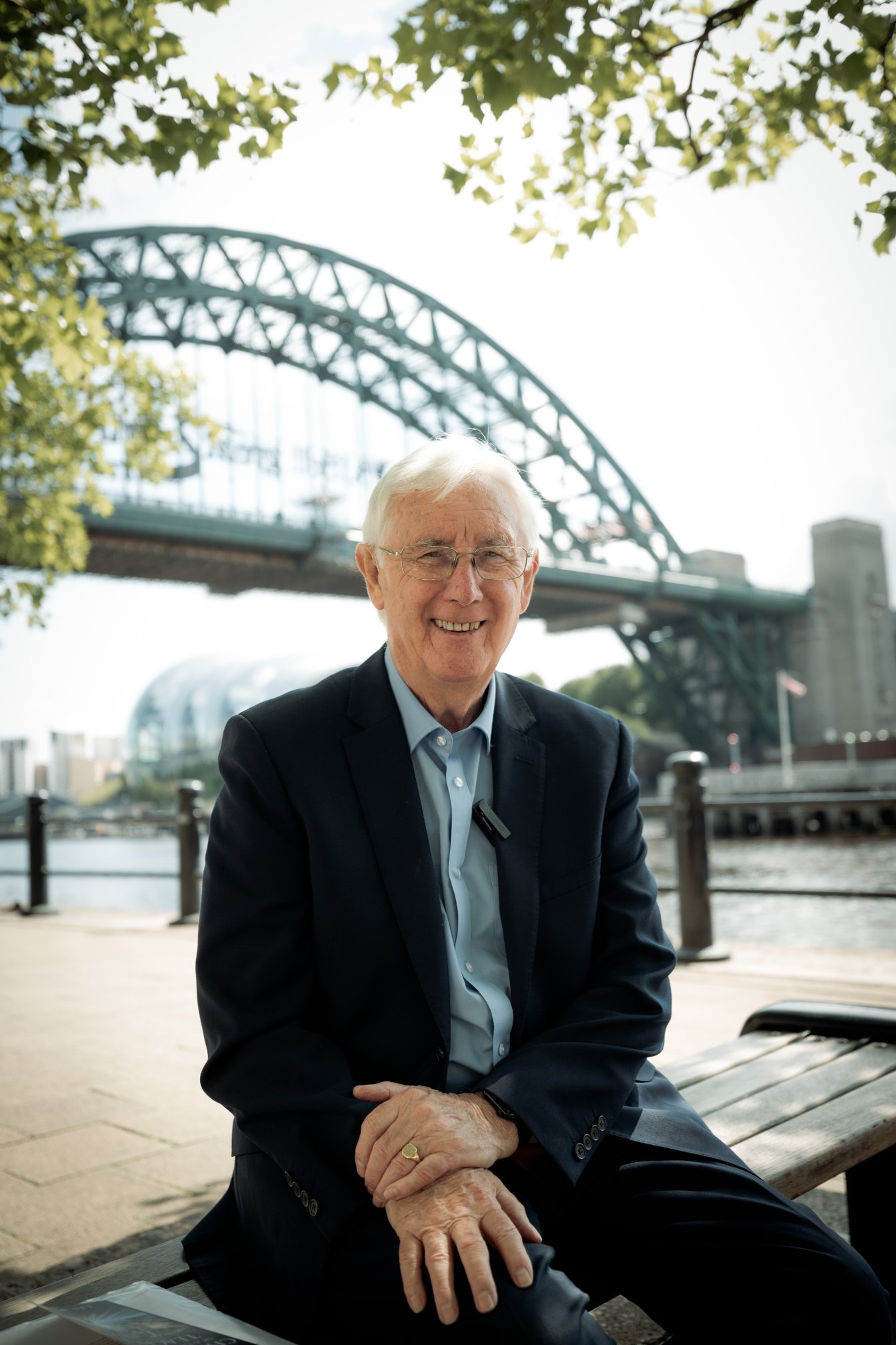 Les Walton CBE seated beside the River Tyne, with the Tyne Bridge behind him